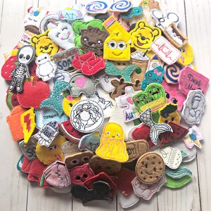 May include: A collection of colorful felt appliques in various shapes and designs. The appliques include cartoon characters, food items, and other whimsical shapes. The appliques are in shades of red, yellow, blue, green, and white.