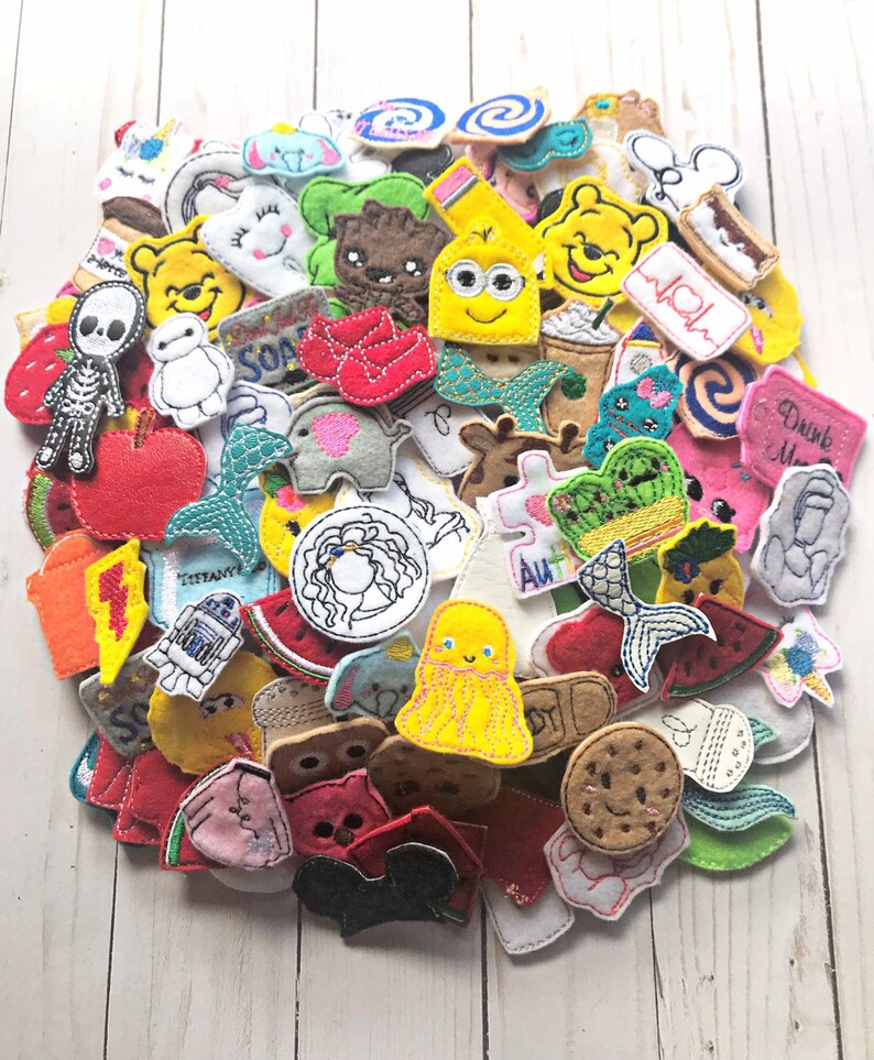 May include: A collection of colorful felt appliques in various shapes and designs. The appliques include cartoon characters, food items, and other whimsical shapes. The appliques are in shades of red, yellow, blue, green, and white.