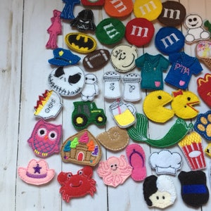 May include: A collection of assorted felt embroidered patches featuring various shapes including characters, food items, vehicles, and symbols. The colorful fabric pieces display detailed stitching and diverse designs, such as a tractor, owl, crab, octopus, and various icons, arranged in a scattered layout.