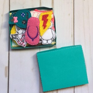 May include: A small, open teal box filled with colorful felt appliques. The appliques include a pink flip-flop, a teal bow, a yellow lightning bolt, a white cartoon character, and a drawing of a person.