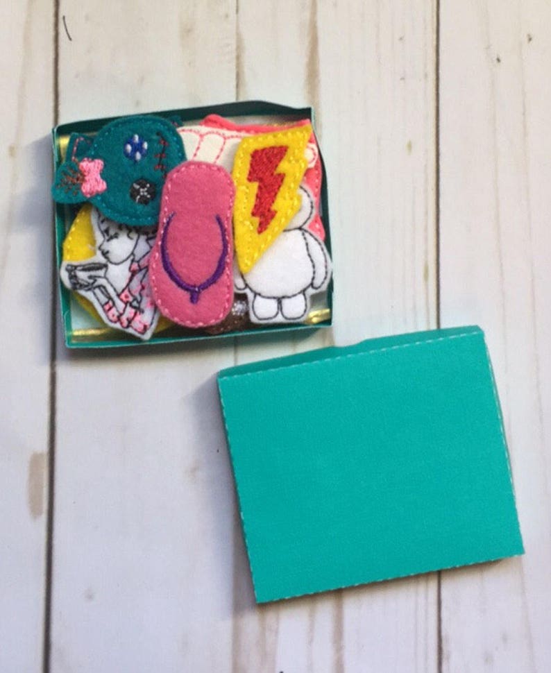 May include: A small, open teal box filled with colorful felt appliques. The appliques include a pink flip-flop, a teal bow, a yellow lightning bolt, a white cartoon character, and a drawing of a person.