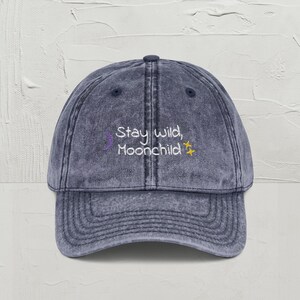 May include: A denim blue baseball cap with a curved brim. The cap features the embroidered text "Stay wild, Moonchild" in white, with a purple crescent moon and a yellow star. The hat has a vintage, worn look.