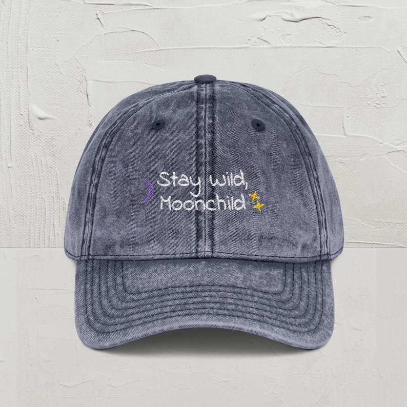 May include: A denim blue baseball cap with a curved brim. The cap features the embroidered text "Stay wild, Moonchild" in white, with a purple crescent moon and a yellow star. The hat has a vintage, worn look.