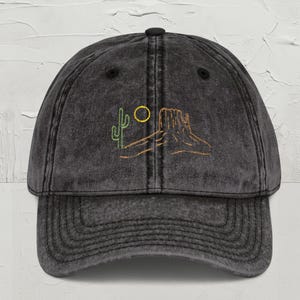 May include: A dark gray baseball cap with a pre-washed, vintage look. The cap features an embroidered desert landscape design in green and gold, including a cactus, sun, and rock formation. The cap has a curved brim and an adjustable strap.