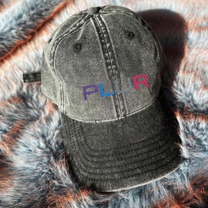 May include: A black distressed denim baseball cap with the word "PLUR" embroidered in rainbow colours.