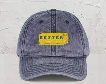 May include: A denim blue baseball cap with a curved brim. The cap features an embroidered yellow rectangle with the word "BUTTER" in blue block letters. The hat has a vintage, worn look.