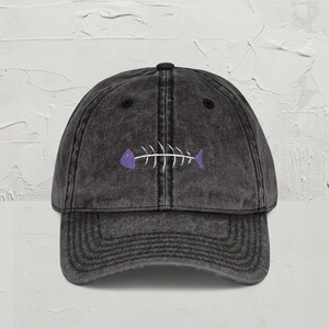 May include: A black, vintage baseball cap with a curved peak. The cap features a white fish skeleton embroidered on the front, with purple accents. The hat has a distressed appearance.