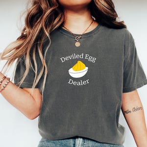 May include: Dark gray t-shirt with the text "Deviled Egg Dealer" above a graphic of a deviled egg. The graphic shows a white bowl with a yellow egg yolk. The shirt is a short-sleeved, crew neck style.