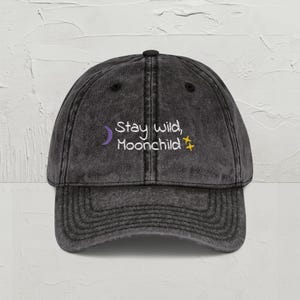May include: A black, vintage-style baseball cap with the embroidered text "Stay wild, Moonchild." The text is in white, with a purple crescent moon and yellow star accents. The cap has a curved brim and adjustable strap.