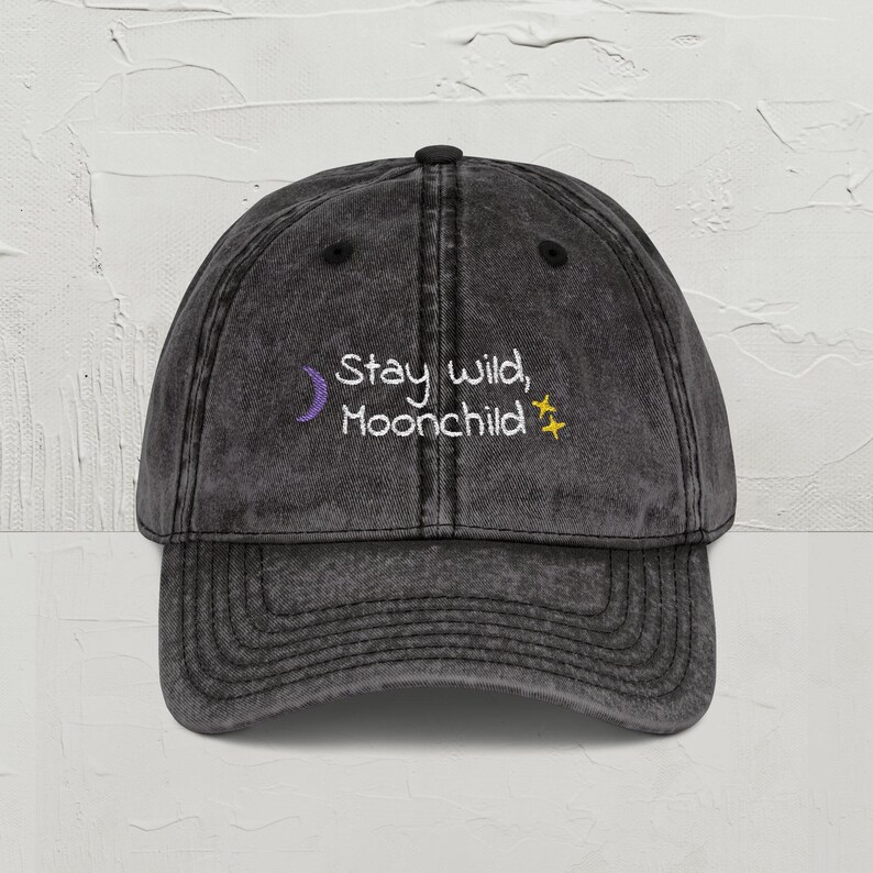May include: A black, vintage-style baseball cap with the embroidered text "Stay wild, Moonchild." The text is in white, with a purple crescent moon and yellow star accents. The cap has a curved brim and adjustable strap.