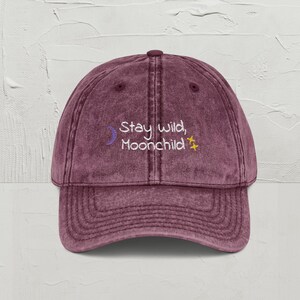 May include: A maroon baseball cap with a curved brim. The cap features the embroidered text "Stay wild, Moonchild" in white, with a crescent moon and star accents. The hat has a vintage, worn look.