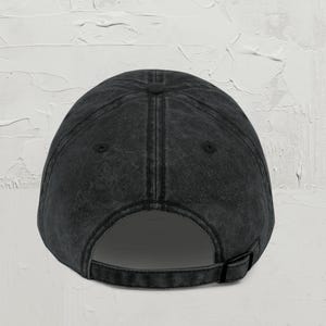 May include: A dark gray baseball cap with a curved brim and adjustable strap. The cap has a vintage, worn look, with visible stitching and a button on top. The back features an adjustable strap for a custom fit.