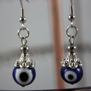 May include: Silver earrings with a blue evil eye charm. The charm has a white center and a black pupil.