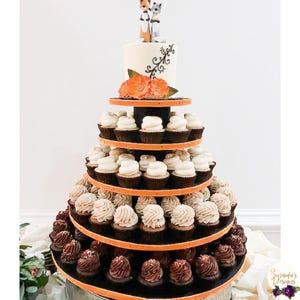 May include: A tiered cupcake stand with a white cake topped with a couple of fox figurines. The stand is decorated with orange and white cupcakes and a black base.