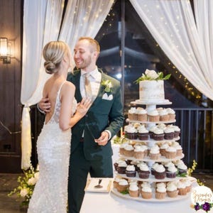 May include: A bride and groom stand in front of a wedding cake with cupcakes. The cake is a semi-naked cake with a white frosting and gold accents. The cupcakes are arranged in a tiered tower with alternating layers of chocolate and vanilla cupcakes. The bride is wearing a white wedding dress and the groom is wearing a dark green suit. The background is a white curtain with twinkling lights.