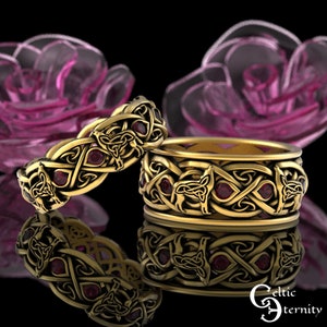 Puede incluir: Dos anillos dorados con intrincados diseños celtas y lobos, acentuados con pequeñas piedras preciosas rojas. Los anillos se exhiben sobre una superficie reflectante, con un fondo rosa borroso. Anillos de boda o compromiso.