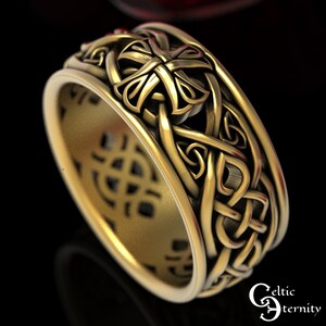 May include: A gold-toned ring featuring an intricate, raised knotwork pattern that wraps around the entire band. The design includes repeating cross-like motifs integrated into the woven metal structure, with a smooth, polished interior surface.