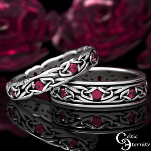 Puede incluir: Dos anillos de plata con diseños de nudos celtas y piedras preciosas rojas incrustadas. Los anillos se muestran sobre una superficie reflectante, con un fondo borroso de tonos rojos. Los anillos son de Celtic Eternity.