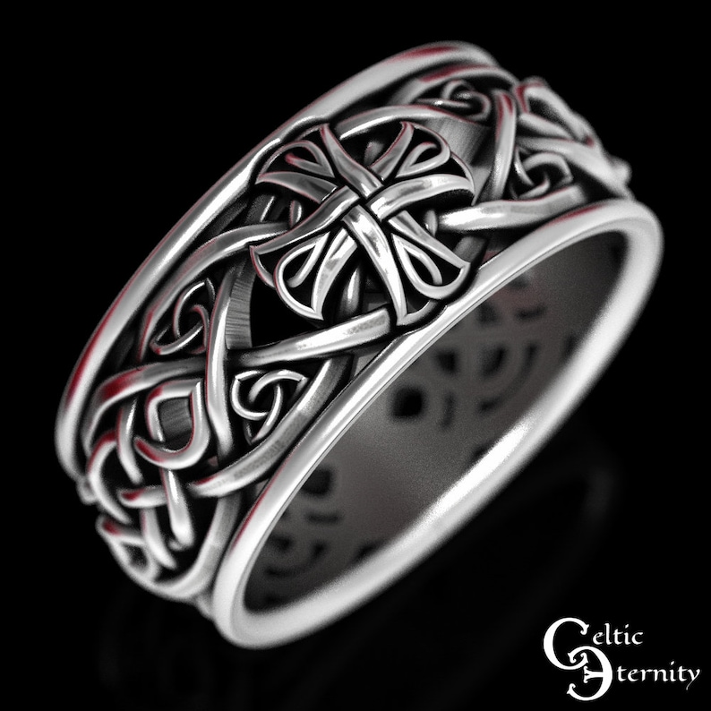 May include: A silver-toned ring featuring an intricate, raised relief pattern of interwoven knots and a central cross motif. The band has a smooth, polished interior surface and a textured, detailed exterior. The text Celtic Eternity is visible.
