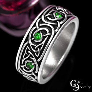 Könnte beinhalten: Ein silberner Ring mit einem keltischen Knotenmuster und drei grünen Edelsteinen. Der Ring ist breit und hat ein detailliertes Design.