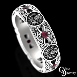 Puede incluir: Un anillo de plata con un nudo celta, tres piedras preciosas rojas y un diseño de cardo. El anillo es un símbolo tradicional de Escocia.