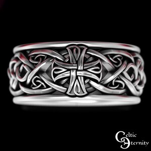 May include: A silver-toned ring featuring a continuous band with an embossed cross motif centered among intricate, interwoven knotwork patterns. The metal surface has a polished finish with darkened recessed areas to highlight the detailed design.