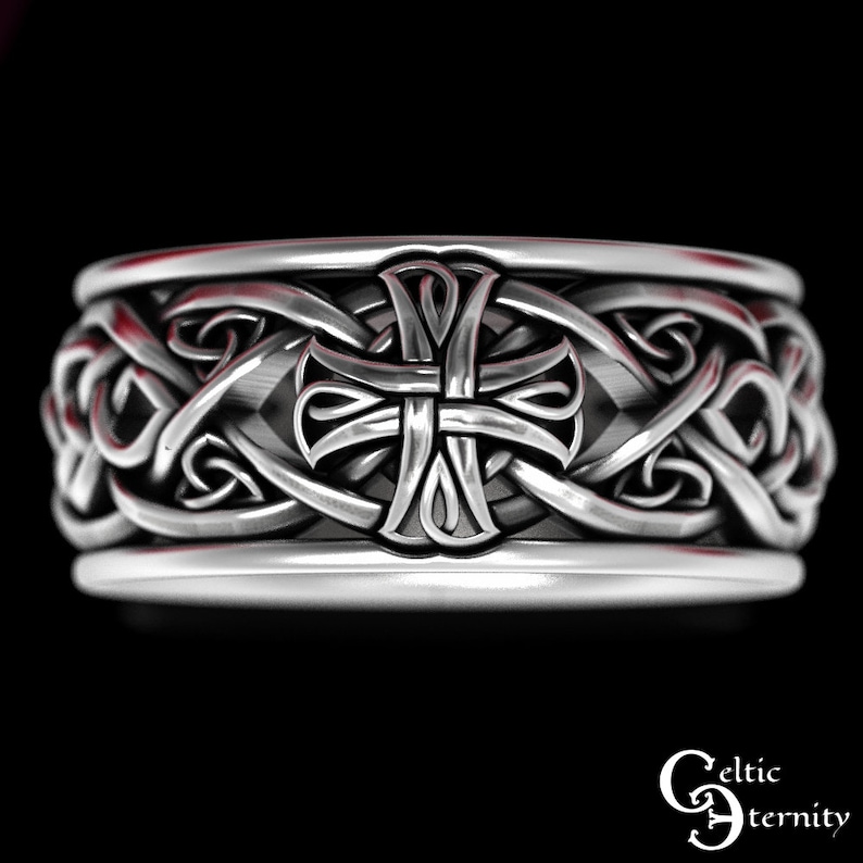 May include: A silver-toned ring featuring a continuous band with an embossed cross motif centered among intricate, interwoven knotwork patterns. The metal surface has a polished finish with darkened recessed areas to highlight the detailed design.
