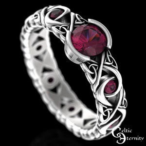 以下が含まれることがあります： 中央に大きな丸い濃い赤色の宝石、両側に小さな丸い濃い赤色の宝石が2つ付いた、銀製のケルト結び目のリング。リングはケルト結び目で複雑にデザインされています。