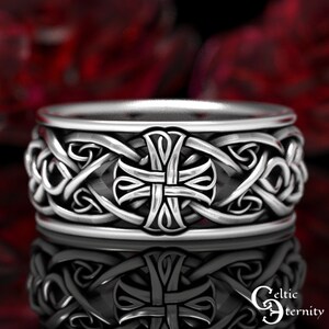 May include: A silver-toned metal ring featuring a continuous band with an embossed cross motif at the center, surrounded by intricate, raised interlacing knotwork patterns. The surface has a polished finish with darkened recessed areas.