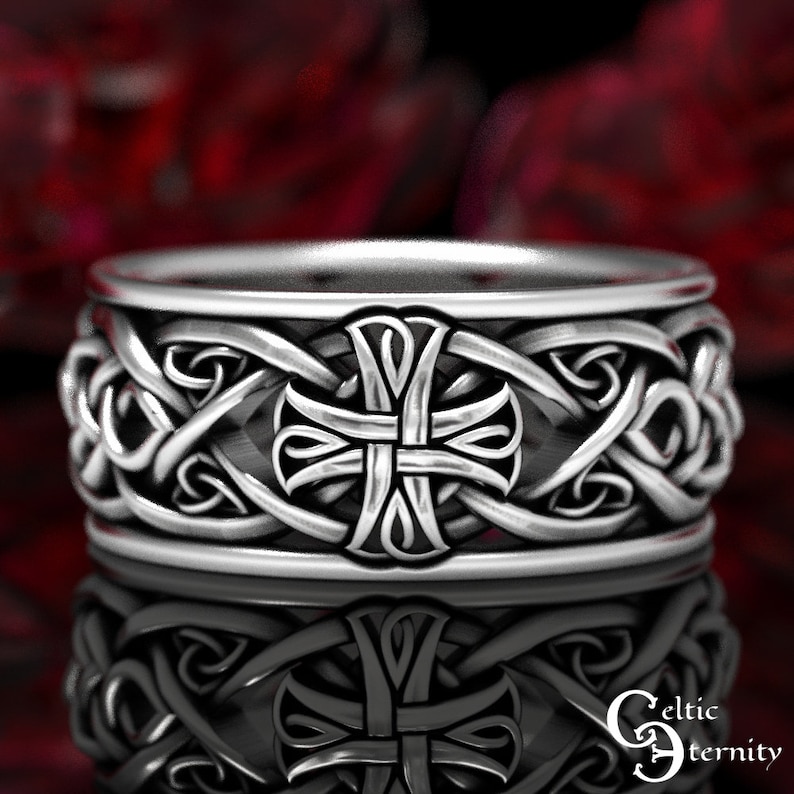 May include: A silver-toned metal ring featuring a continuous band with an embossed cross motif at the center, surrounded by intricate, raised interlacing knotwork patterns. The surface has a polished finish with darkened recessed areas.