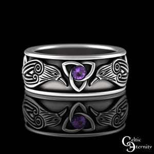 May include: A silver-colored ring with a Celtic design. The ring features a central purple gemstone within a triquetra knot, flanked by stylized bird or raven motifs. The ring's band is wide and has a polished finish.