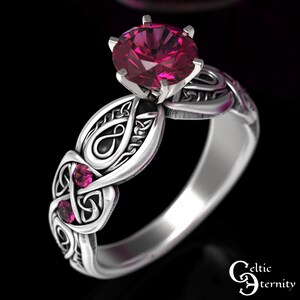 Puede incluir: Un anillo de plata con un nudo celta adornado con una gran piedra preciosa rosa en el centro y dos piedras rosas más pequeñas en la banda. El anillo está diseñado con intrincados nudos celtas.