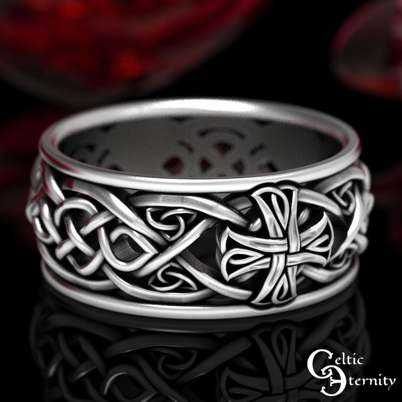 May include: A silver-toned ring featuring an intricate, continuous Celtic knotwork pattern that wraps around the entire band. The design incorporates a prominent cross motif integrated into the metalwork, with a polished finish and dark recessed areas.