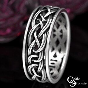 May include: A silver-toned metal ring featuring an intricate, raised Celtic knotwork pattern around the exterior band. The inner surface of the ring displays a repeating diamond-shaped cutout design. The text Celtic Eternity is visible.