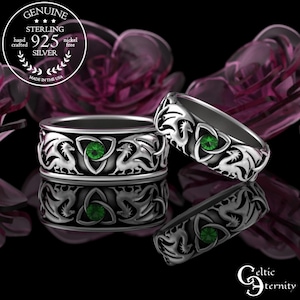 Könnte beinhalten: Zwei Ringe aus Sterlingsilber mit einem wiederkehrenden Drachenmotiv und einem zentralen grünen Edelstein in einem Triquetra-Knotendesign. Das Metall hat eine polierte Oberfläche mit abgedunkelten vertieften Bereichen, um die Drachengravuren zu betonen.