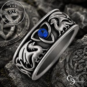 May include: A sterling silver ring with a dragon design. The ring features two dragons on the sides and a blue gemstone in the center, set within a Celtic knot. The ring is marked "925 SILVER" and "MADE IN THE USA".