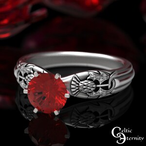 Puede incluir: Un anillo de plata con una piedra preciosa roja engastada en el centro. El anillo presenta un diseño de nudo celta con un cardo en un lado y una flor de lis en el otro.