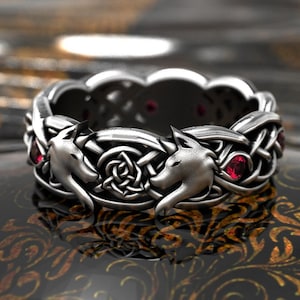 May include: A silver ring featuring two wolf heads facing each other, with a Celtic knot design in the center. The ring has a braided band and is accented with small red gemstones. The ring is displayed on a reflective surface.
