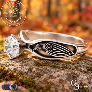 May include: A sterling silver ring featuring a round, clear faceted stone set in a six-prong setting. The band displays an engraved bird motif with stylized wing details. A circular seal in the corner reads Genuine Sterling 925 Silver.