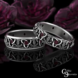 Könnte beinhalten: Zwei silberne Ringe mit keltischen Knoten, roten Edelsteinen und Distelmotiven. Die Ringe sind ineinander verschlungen und haben eine polierte Oberfläche.