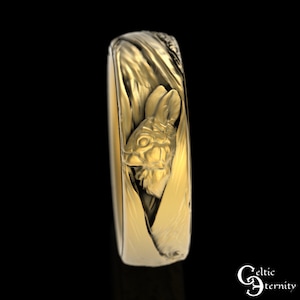 May include: A gold band ring with a detailed, three-dimensional rabbit design. The rabbit's head is peeking out from behind a textured, swirling pattern.
