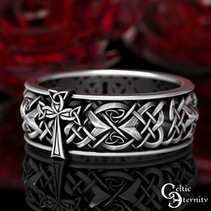 May include: A silver ring with a Celtic knot design and a cross in the center. The ring is on a red background.