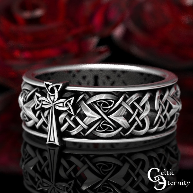 May include: A silver ring with a Celtic knot design and a cross in the center. The ring is on a red background.
