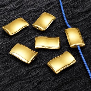 Può includere: Una collezione di sette perline rettangolari color oro con una superficie liscia e curva. Ogni perlina presenta un piccolo foro alle estremità per l'infilatura. Una perlina è infilata su un sottile cordino blu, le altre sono sparse.