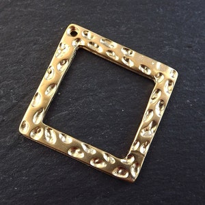 Puede incluir: Colgante de marco cuadrado dorado con superficie texturizada. El marco tiene un agujero en la parte superior para colgar.