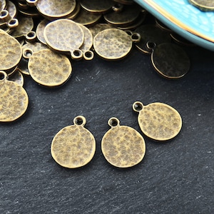 以下が含まれることがあります： アンティークゴールドの金属製チャームの山をクローズアップした写真。チャームは丸型で、ハンマーで叩いたような質感があります。手前に4つのチャームが、山から離れて置かれています。