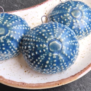 Pode incluir: Três ornamentos de cerâmica azul com salpicos brancos e uma argola de metal para pendurar, apresentados num prato salpicado com borda castanha. Os ornamentos têm um design de ouriço-do-mar.