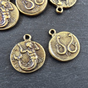 May include: Two gold-toned metal charms featuring zodiac signs. One charm depicts a mermaid with a fish tail and a bow and arrow, with the word "SAGITTARIUS" inscribed around the edge. The other charm depicts two snakes intertwined, with the word "SERPENS" inscribed around the edge.