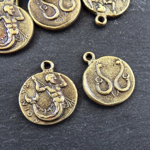 May include: Two gold-toned metal charms featuring zodiac signs. One charm depicts a mermaid with the word "CAPRICORN" inscribed around the edge. The other charm depicts two snakes intertwined with the word "SERPENS" inscribed around the edge.