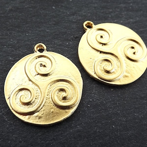 Pode incluir: Dois pingentes redondos dourados com um design espiral em relevo. Cada pingente tem uma pequena argola na parte superior para prender a uma corrente ou outro componente de joalheria. Os pingentes estão em uma superfície escura.